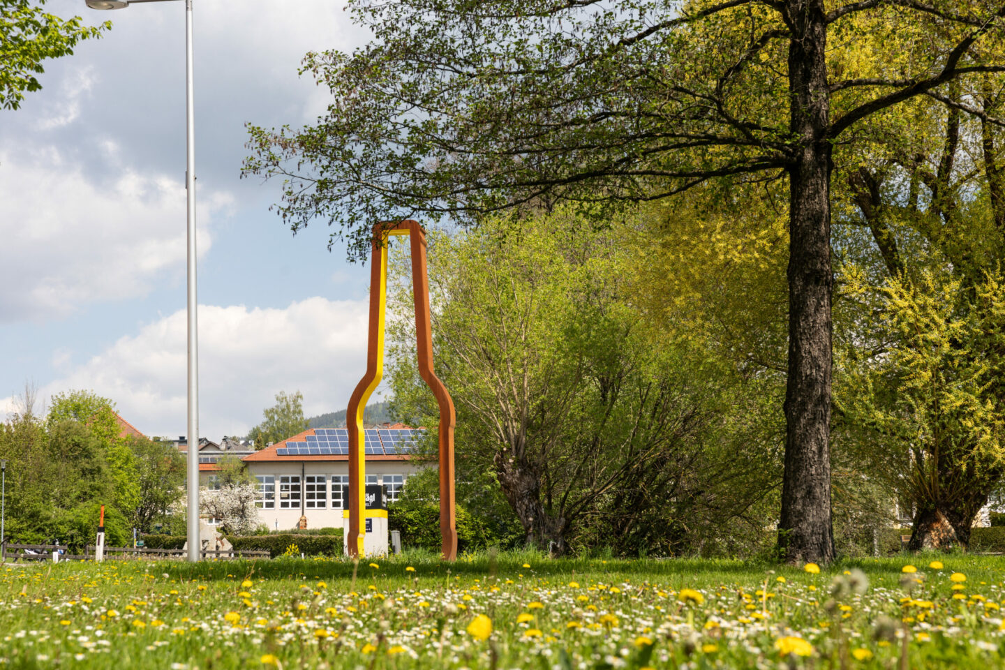 Das Bild zeigt eine Grasfläche mit Löwenzahn im Vordergrund, einer hohen Metallskulptur einer Bierflasche, in der Mitte und Bäumen mit grünem Laub, die das Gebiet umgeben.